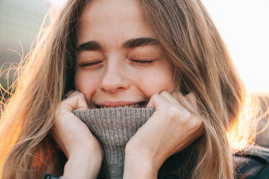 cheerful young woman with eyes closed covering mouth with turtleneck on street, engaging in mindfulness practice for self-care