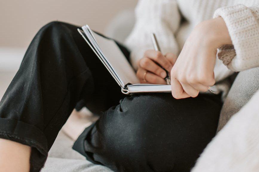 crop young woman writing schedule in diary on sofa