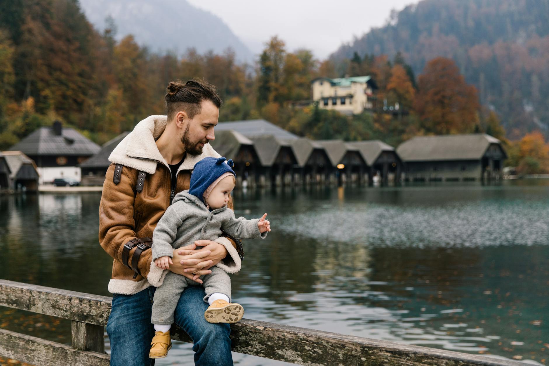 man in brown jacket and blue denim jeans sitting on brown wooden bench beside river during, evoking: natural cycles, Wheel of the Year, seasonal traditions, Sabbats, and holidays.