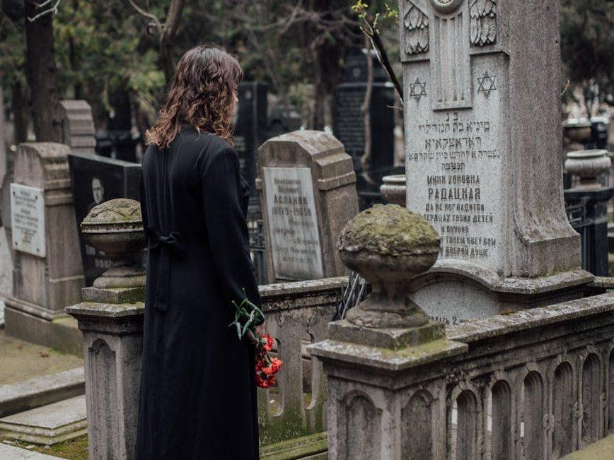 woman in black coat standing in front of a tombstone with flowers