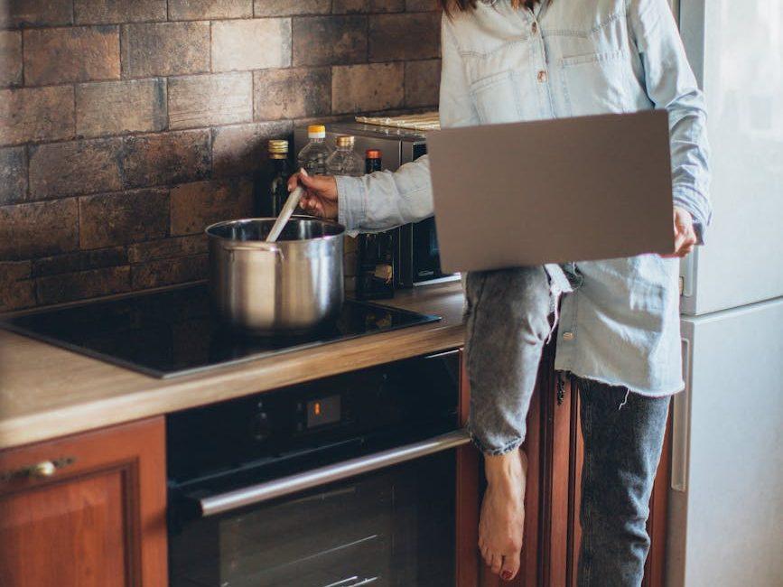 woman in blue denim jacket holding laptop while cooking, illustrating: Samhain, Harvest Season, Stew Recipe, Nourishing Dish, and a Samhain Feast