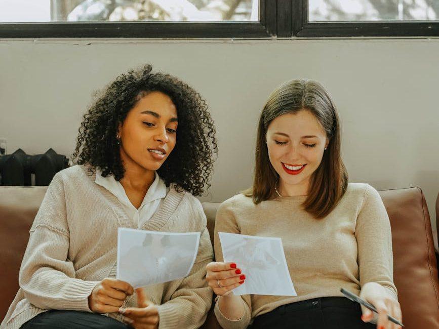 a pair of women sitting on couch holding papers