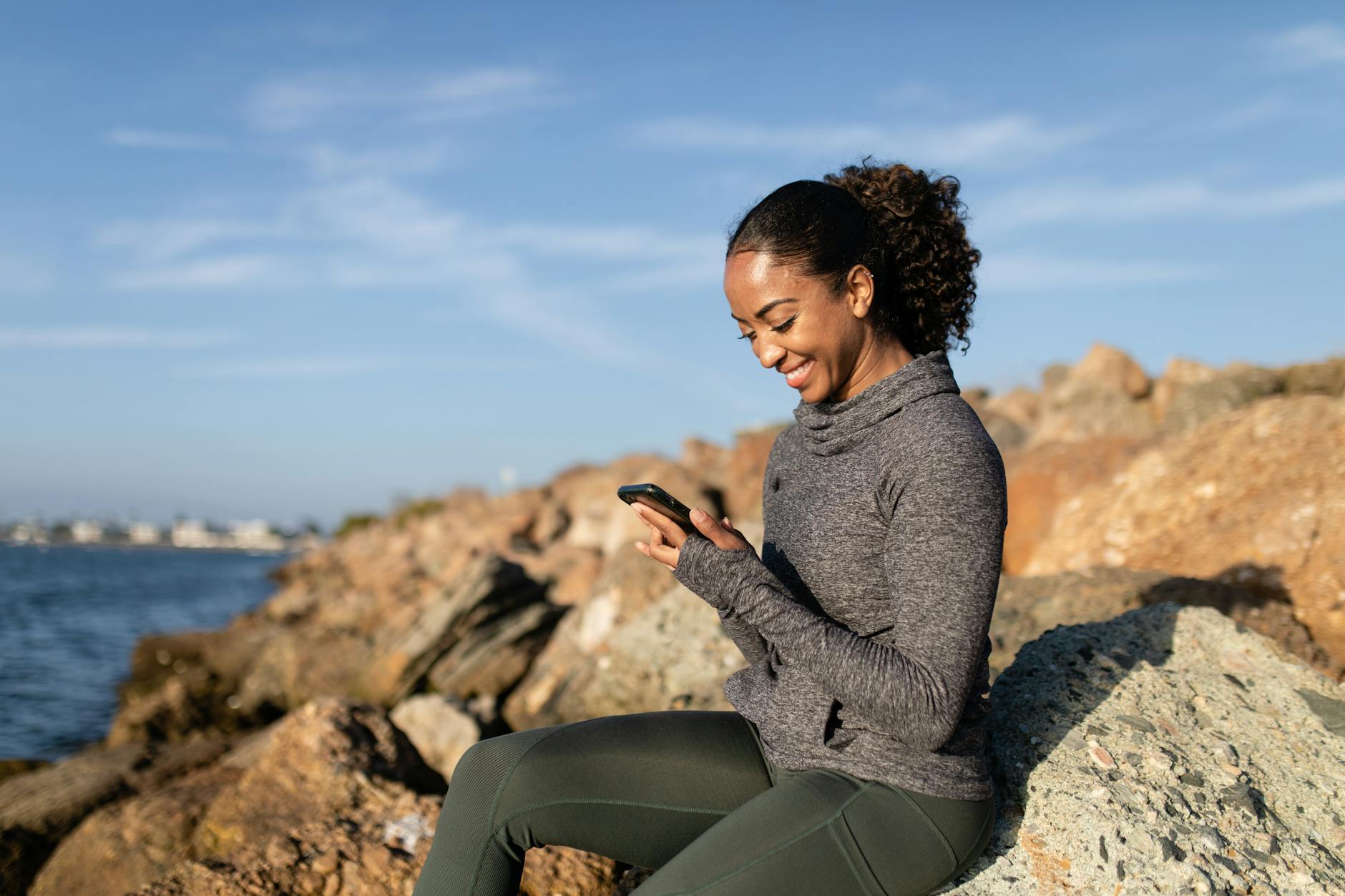 a happy woman using her cellphone while sitting on a rock, enjoying the benefits of her Onesce Insider personal development membership