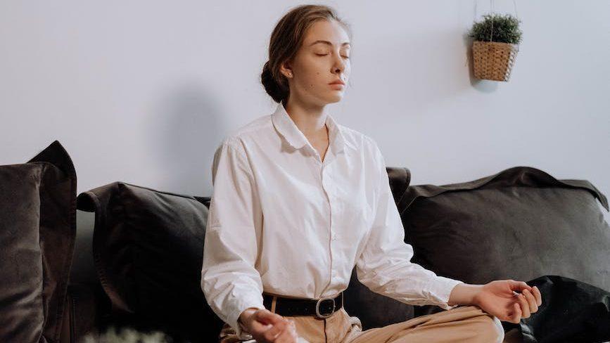 woman in white button up long sleeve shirt sitting on black couch in lotus position, considering personalized meditation, meditation goals, stress reduction, daily mindfulness, and meditation practice