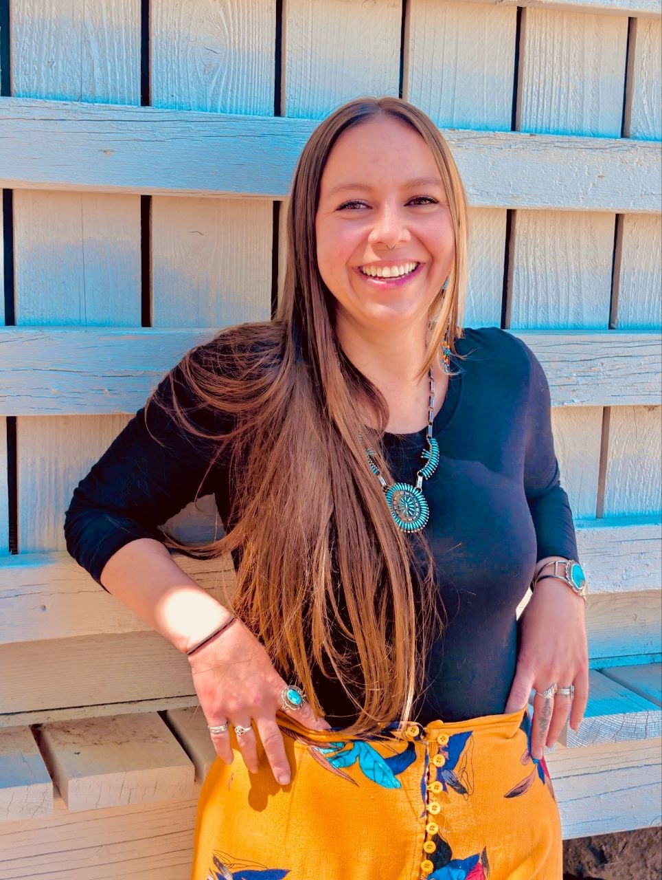 A smiling woman with long hair poses against a wooden backdrop, wearing a black top and a patterned skirt. She has several rings and a necklace, exuding a cheerful and approachable demeanor