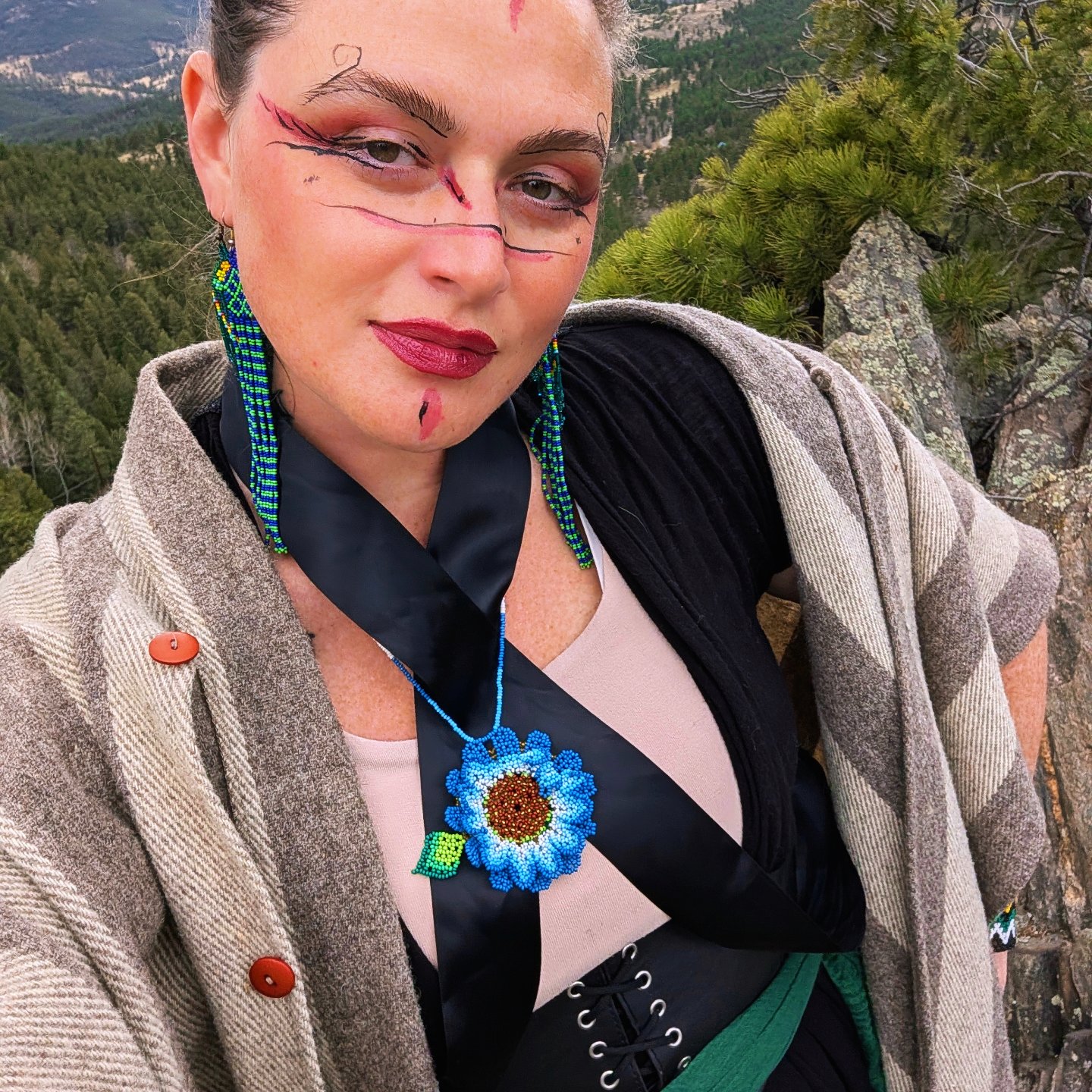 A woman poses outdoors with a scenic mountainous background, wearing a unique outfit featuring a black ribbon, a blue flower necklace, and intricate facial makeup; she has long, colorful earrings and a patterned shawl draped over her shoulders.